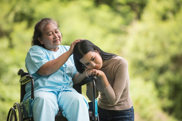 Elderly woman in a wheelchair sharing a comforting moment with a younger woman outdoors, promoting disability inclusion and caregiving support.